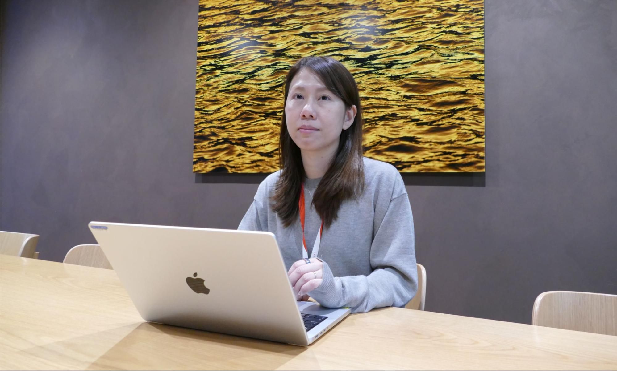 A woman sits at a wooden table with a silver laptop, looking thoughtfully aside.
