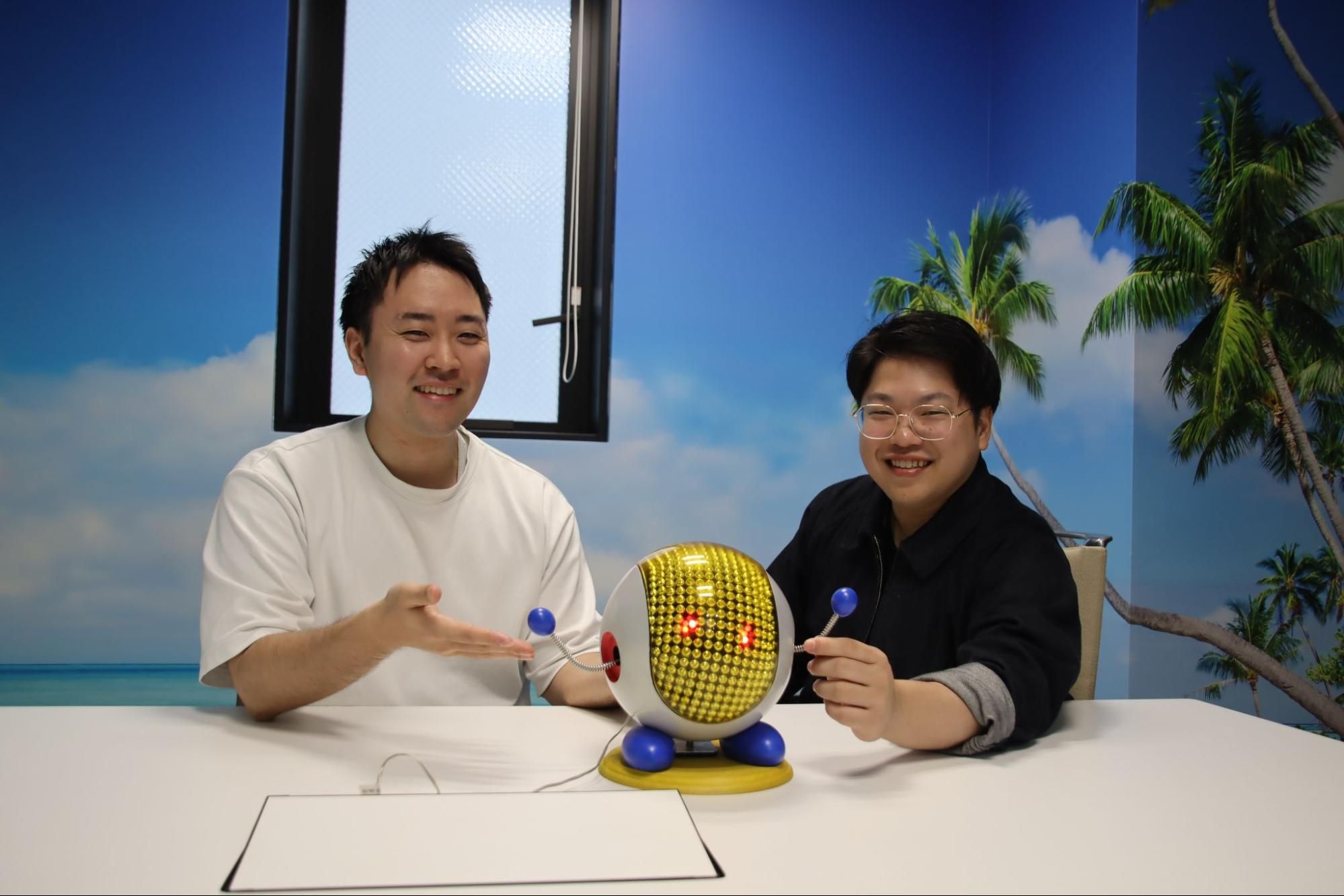 Two men interact with a small, whimsical robot holding a lollipop at a table.