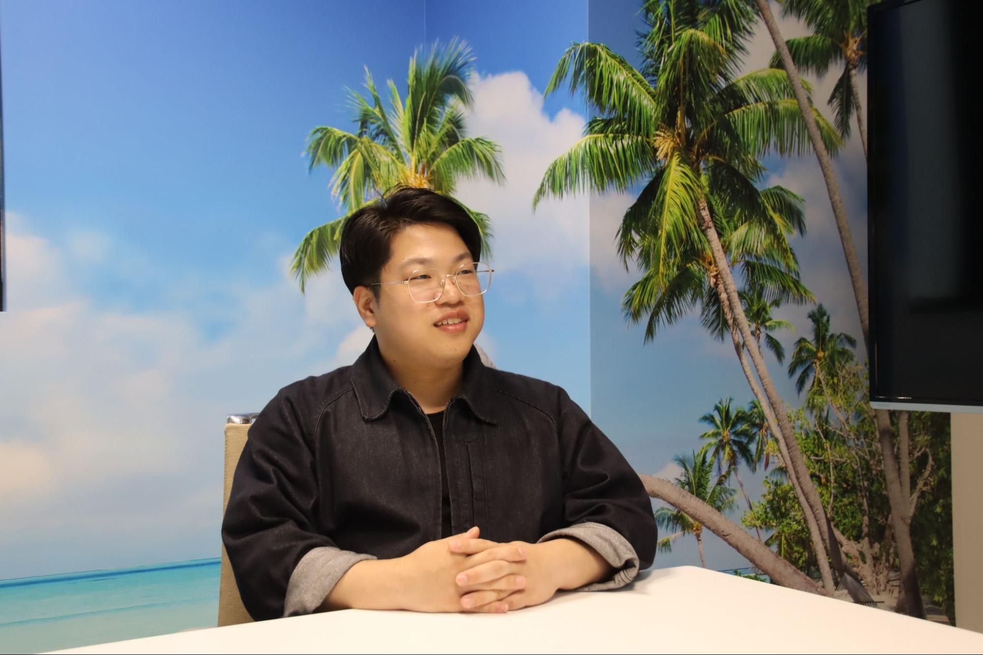 Smiling man in dark shirt at table, tropical beach and palm trees in background.